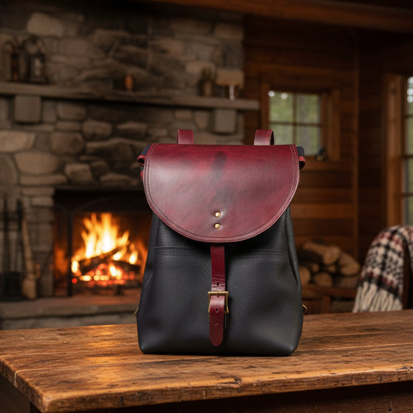 A black leather sbackpack with a large curved front flap, a matching leather strap and buckle, and a black leather body, laid flat on a light fabric surface.