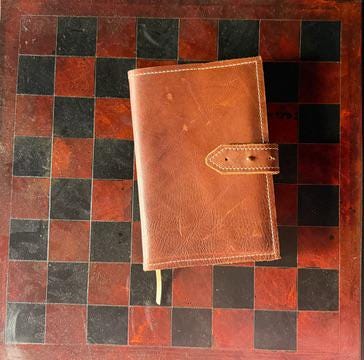A brown leather notebook or planner cover with a strap closure, placed on a red and black checkered board background.