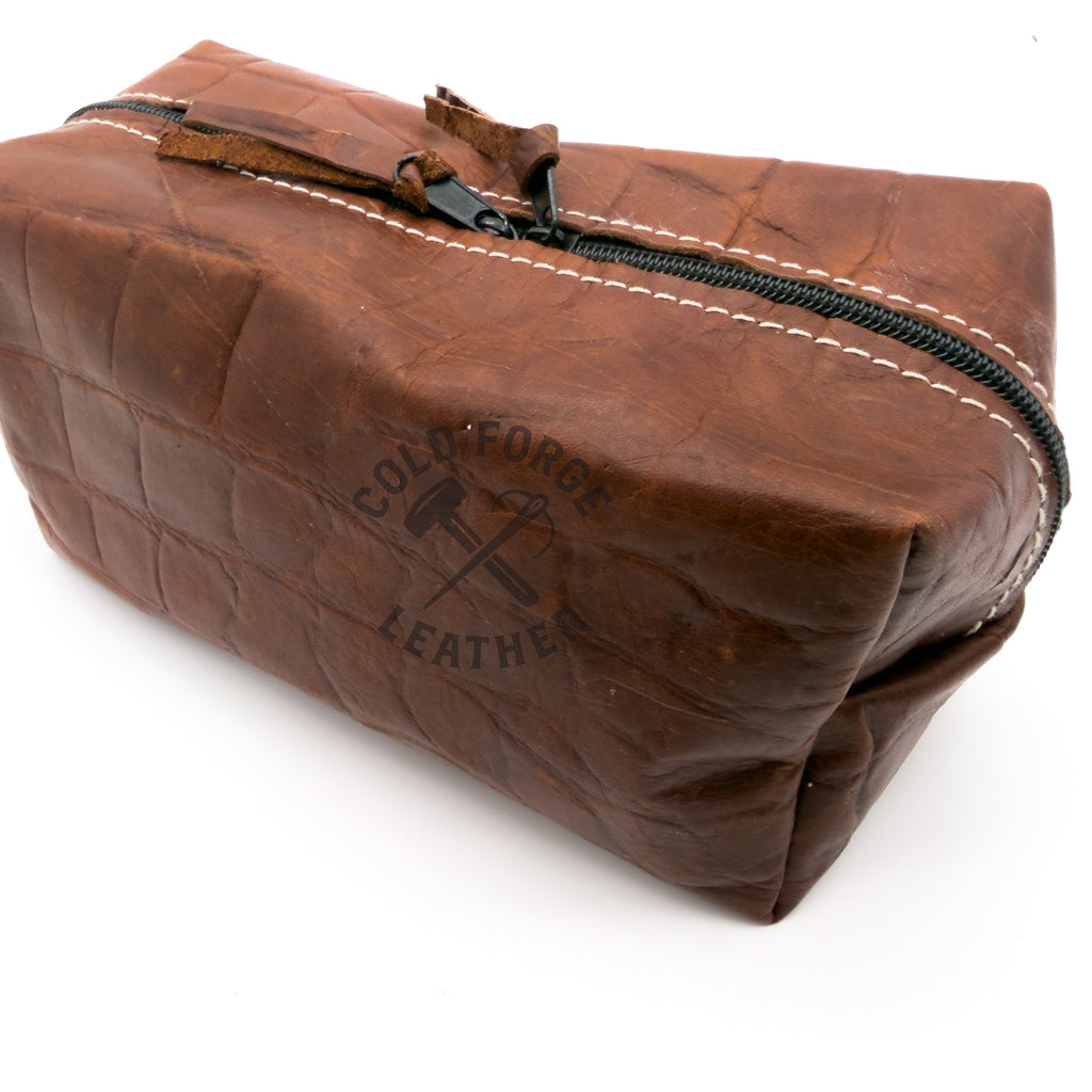 A brown textured leather zippered pouch with white contrast stitching and a black zipper, set against a plain white background.