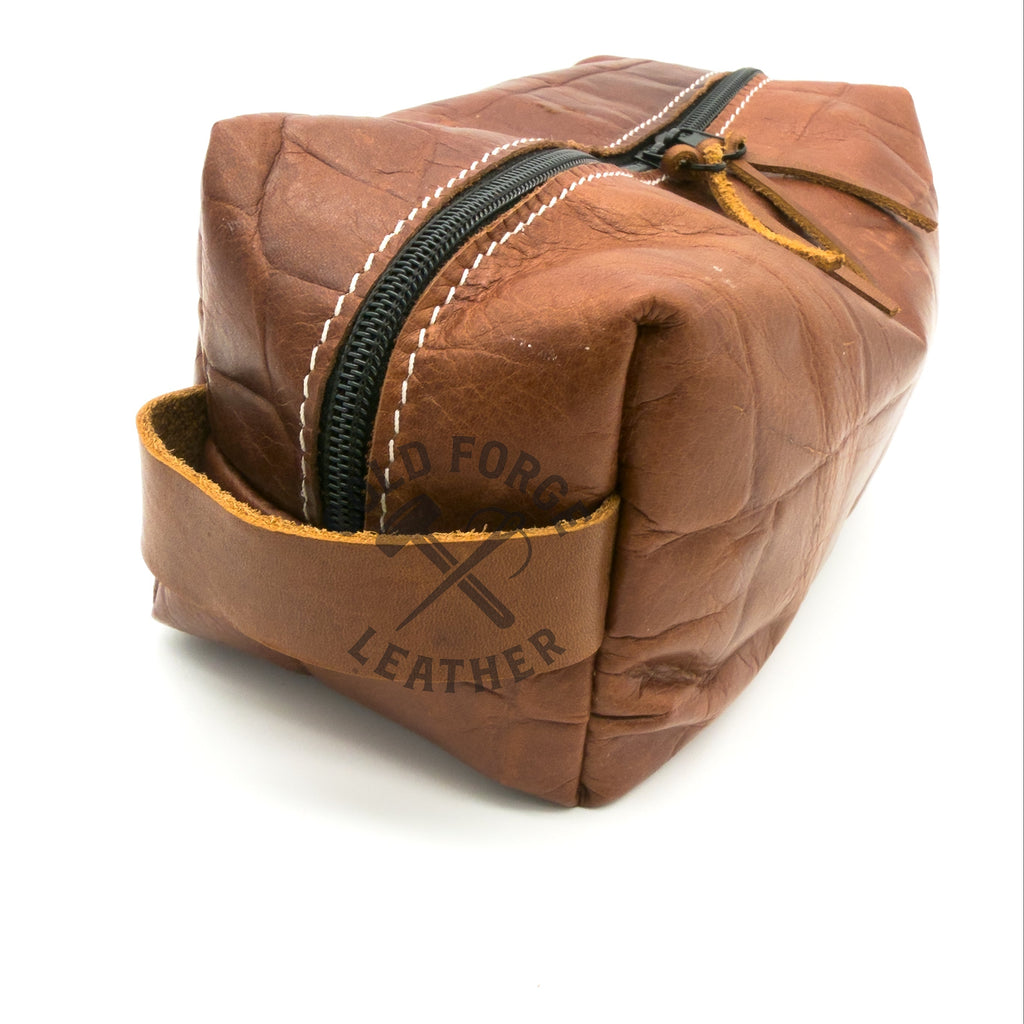 A brown textured leather zippered pouch with white contrast stitching and a black zipper, shown on a white background with a leather stamp on the front pocket.
