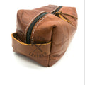 A brown textured leather zippered pouch with white contrast stitching and a black zipper, shown on a white background with a leather stamp on the front pocket.