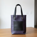 A dark purple leather tote bag with black leather handles and a black front pocket.
