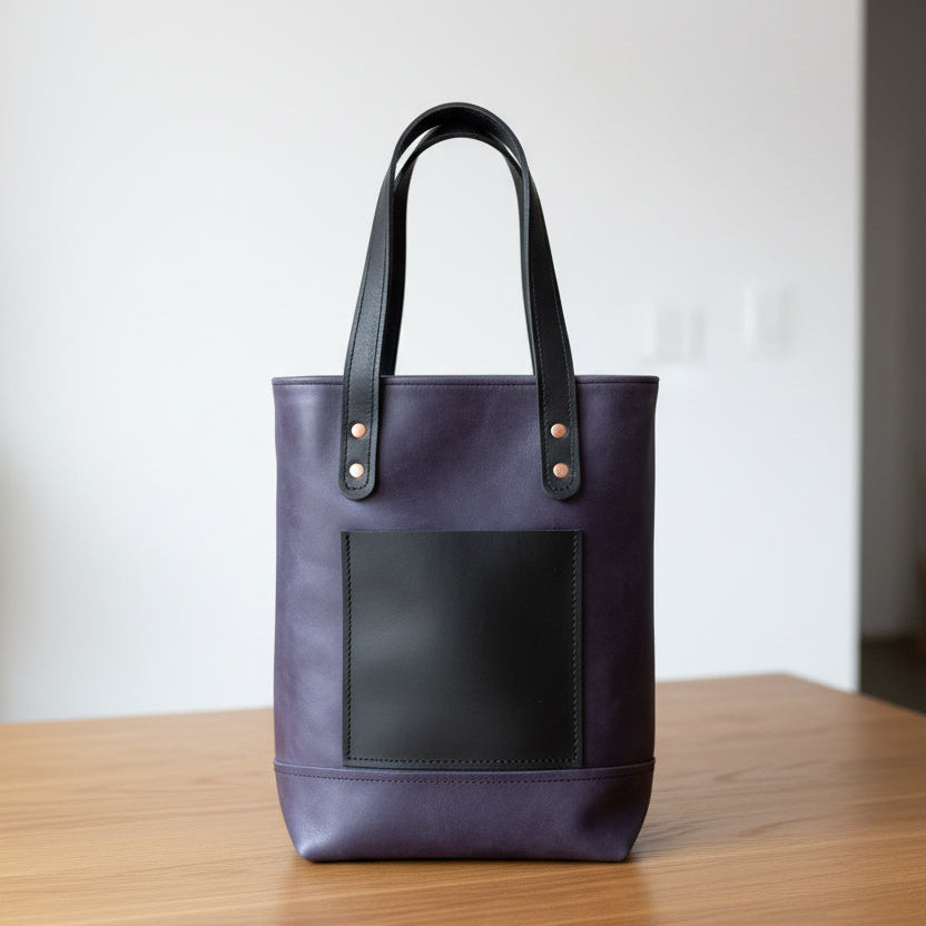 A dark purple leather tote bag with black leather handles and a black front pocket.