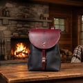 A black leather sbackpack with a large curved front flap, a matching leather strap and buckle, and a black leather body, laid flat on a light fabric surface.