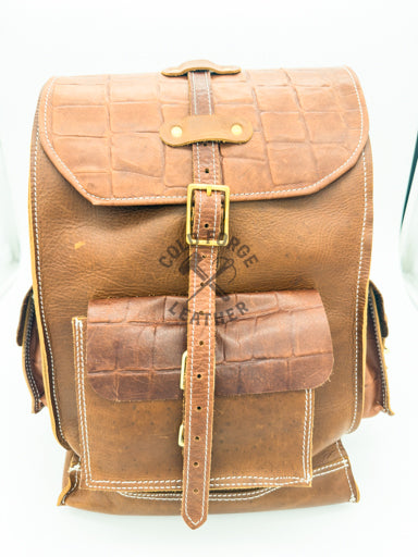 A brown leather backpack with crocodile-embossed textures, gold-tone buckles, and white contrast stitching, shown against a plain white background.