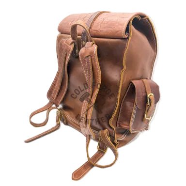 A brown leather backpack with a textured crocodile pattern on the top flap, adjustable shoulder straps, and a side pocket with a brass buckle, photographed on a white studio background.