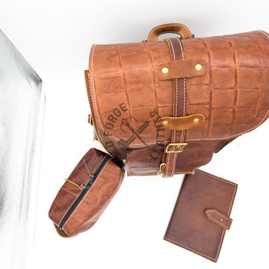 A brown leather tool belt with a matching Dopp kit and journal, displayed on a reflective white surface. The tool belt features a crocodile-embossed texture, brass buckle details.