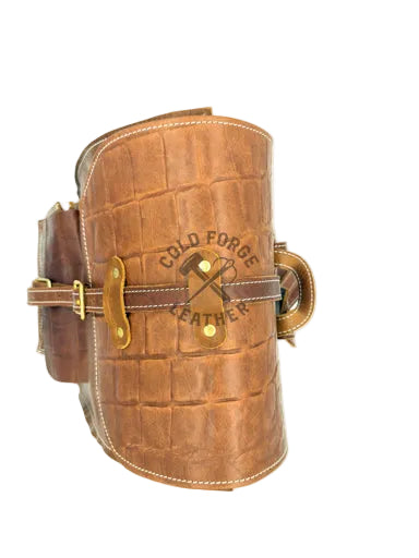 A brown leather shield with an embossed crocodile pattern, featuring brass hardware, a front buckle strap, and a central logo that reads 'COLD FORGE EXPLORER' with a crossed hammer and pickaxe design.