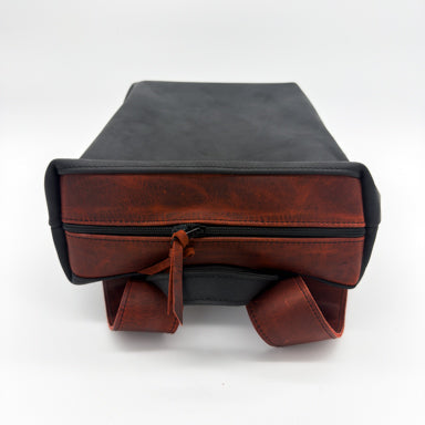A minimalist leather backpack shown from above on a white studio background. The product has a black body and a distressed reddish closure section with a matching redish leather straps and zipper pull.