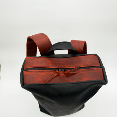 A minimalist leather backpack with a two-tone design featuring a black body and reddish top panels and straps, shown on a white background.