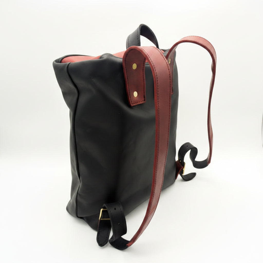 A black leather backpack shown from a side angle on a white background, featuring reddish leather shoulder straps and a black adjustable buckle strap.