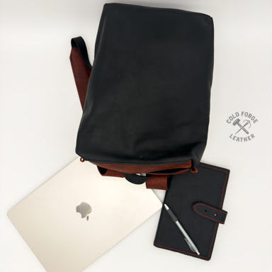 A black leather backpack with reddish leather trim and strap, styled in a flat-lay with a silver laptop, a black leather notebook, and a pen on a white background.