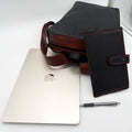 A black leather backpack and notebook cover a, displayed next to a silver laptop, pen, and a white textured background. A 'Cold Forge Leather' logo is visible on the right side.