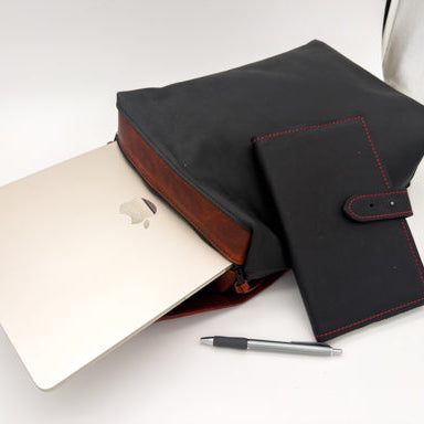 A black leather backpack with reddish leather trim, shown holding a silver laptop. A matching black notebook with red stitching and a pen are placed beside it on a white surface.