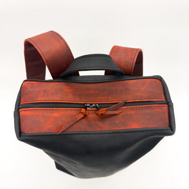 A handmade leather backpack with a two-tone design of reddish trim and black body, featuring a top zipper, top handle, and shoulder straps, shown on a white background.