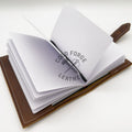 An open brown leather notebook or journal with white dotted pages, shown on a clean white background. A black ribbon is tucked inside, and the pages are slightly fanned out.
