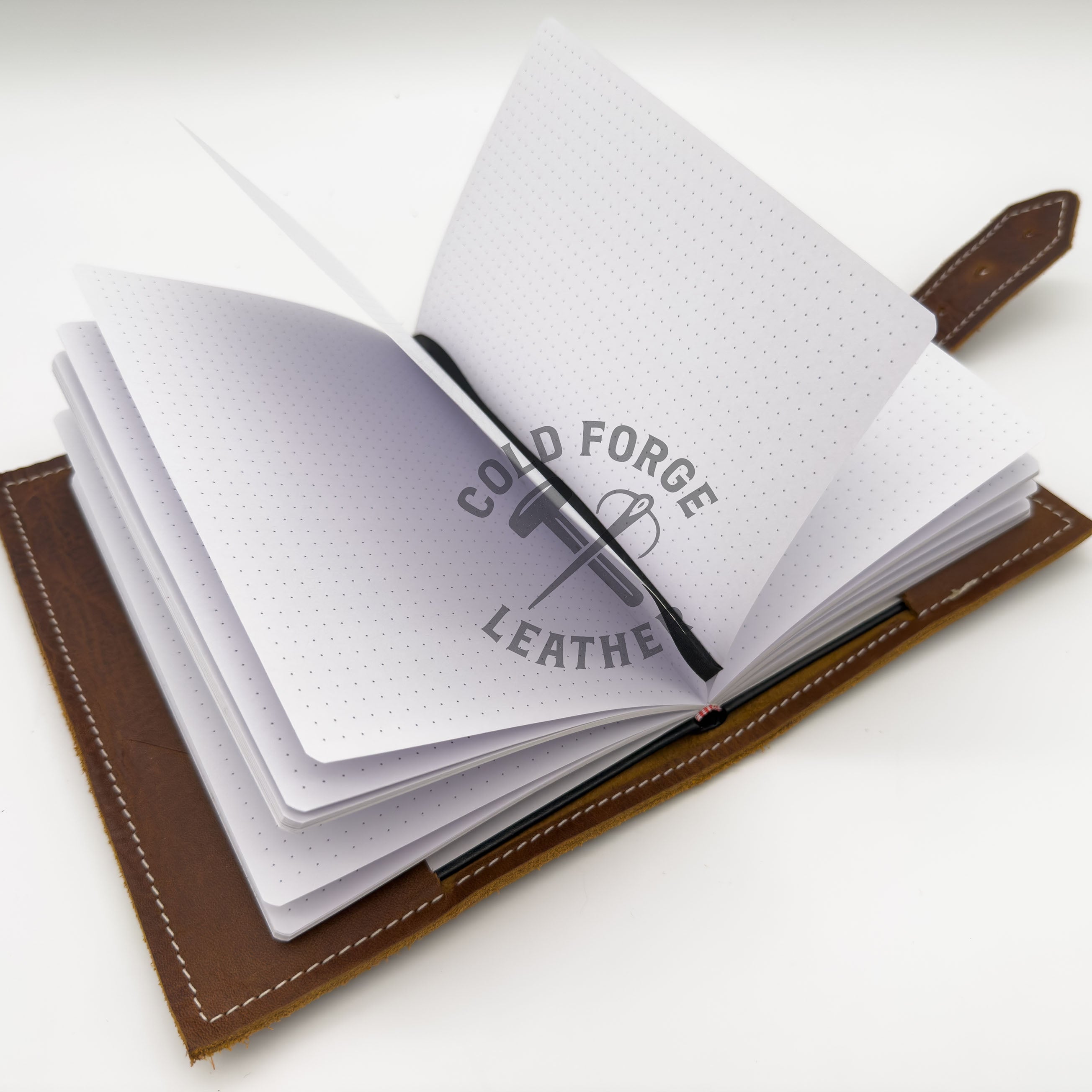 An open brown leather notebook or journal with white dotted pages, shown on a clean white background. A black ribbon is tucked inside, and the pages are slightly fanned out.