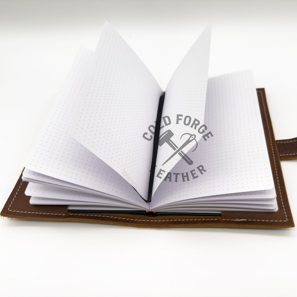 An open brown leather notebook or journal with white dotted pages, shown on a clean white background. A black ribbon is tucked inside, and the pages are slightly fanned out.