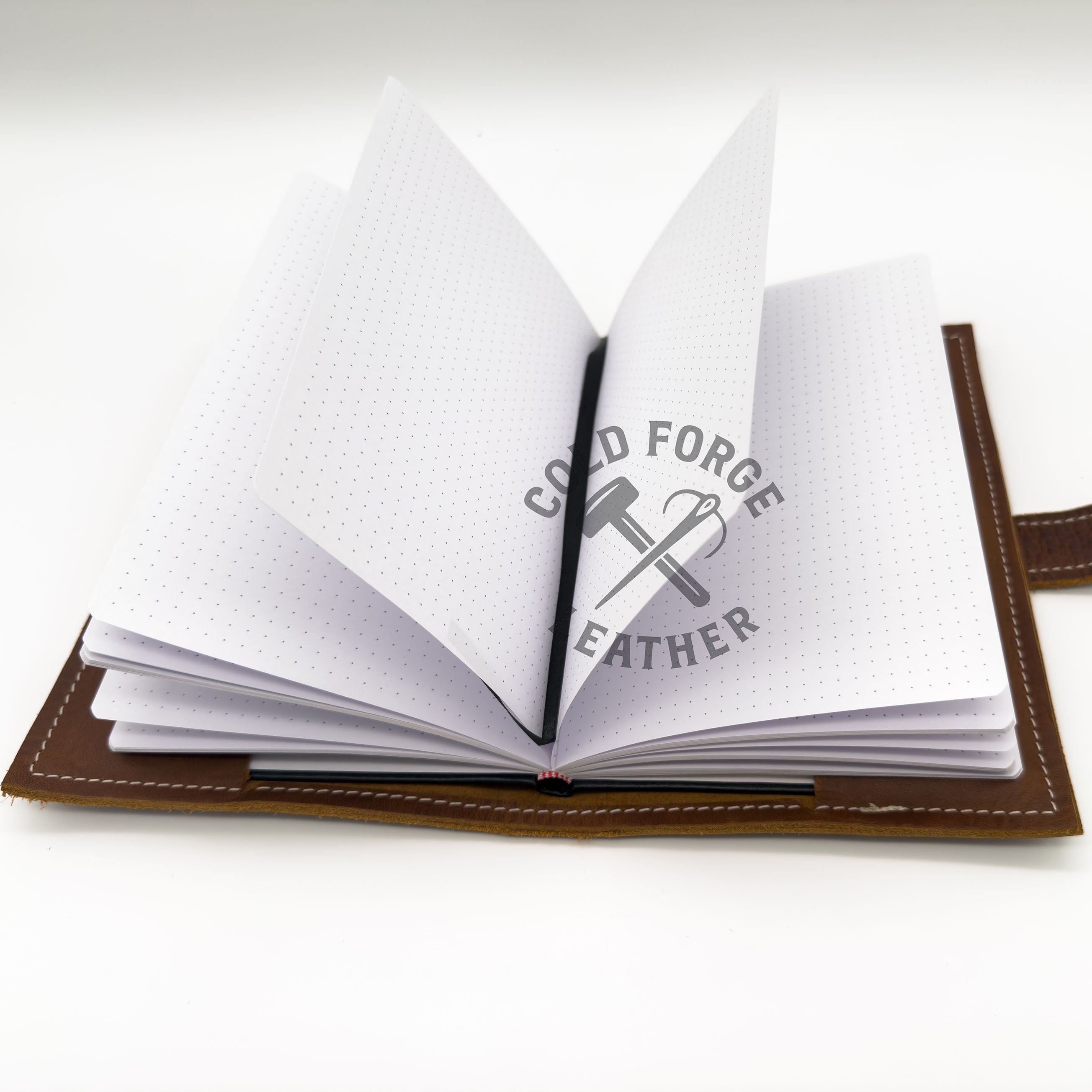 An open brown leather notebook or journal with white dotted pages, shown on a clean white background. A black ribbon is tucked inside, and the pages are slightly fanned out.