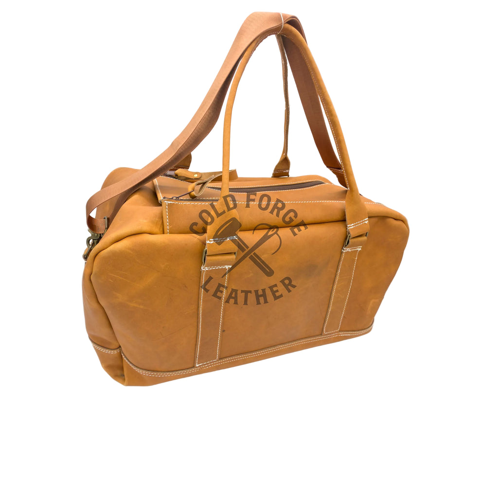 A tan leather duffel bag with a top handle, shoulder strap, and a front zipper pocket featuring a hammer-and-wrench graphic that reads 'COLD FORGE LEATHER'.