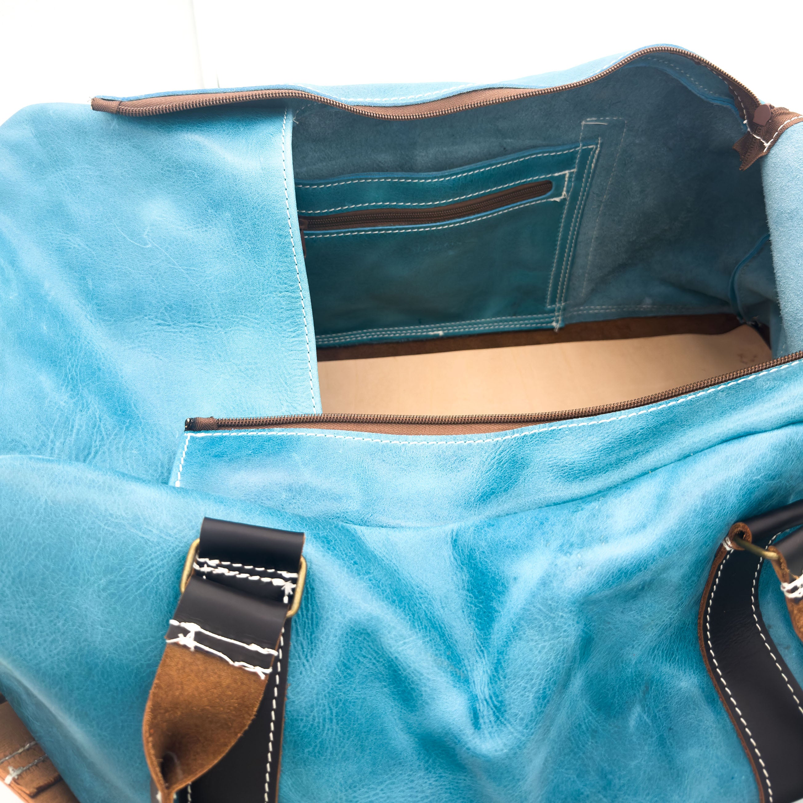 Close-up of a large turquoise blue leather duffel bag showing the interior, zipper, pocket, and dark brown leather strap details.
